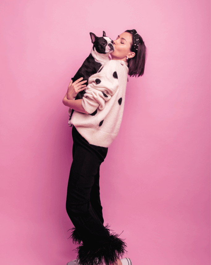 Woman in polka dot sweater holding and kissing a Boston Terrier puppy against a pink background.